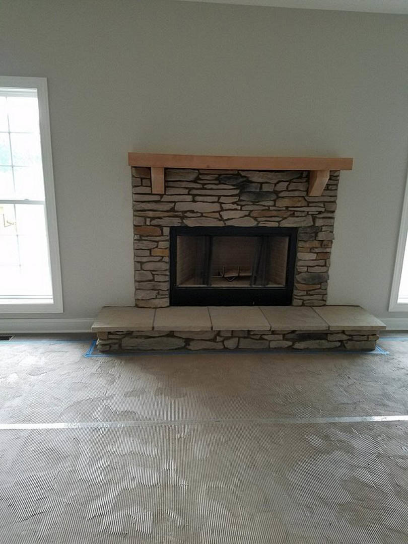 Stone fireplace with hearth and fire screen set against a textured stone wall, adjacent to a window with a white frame and wooden bench, hardwood flooring throughout the room.