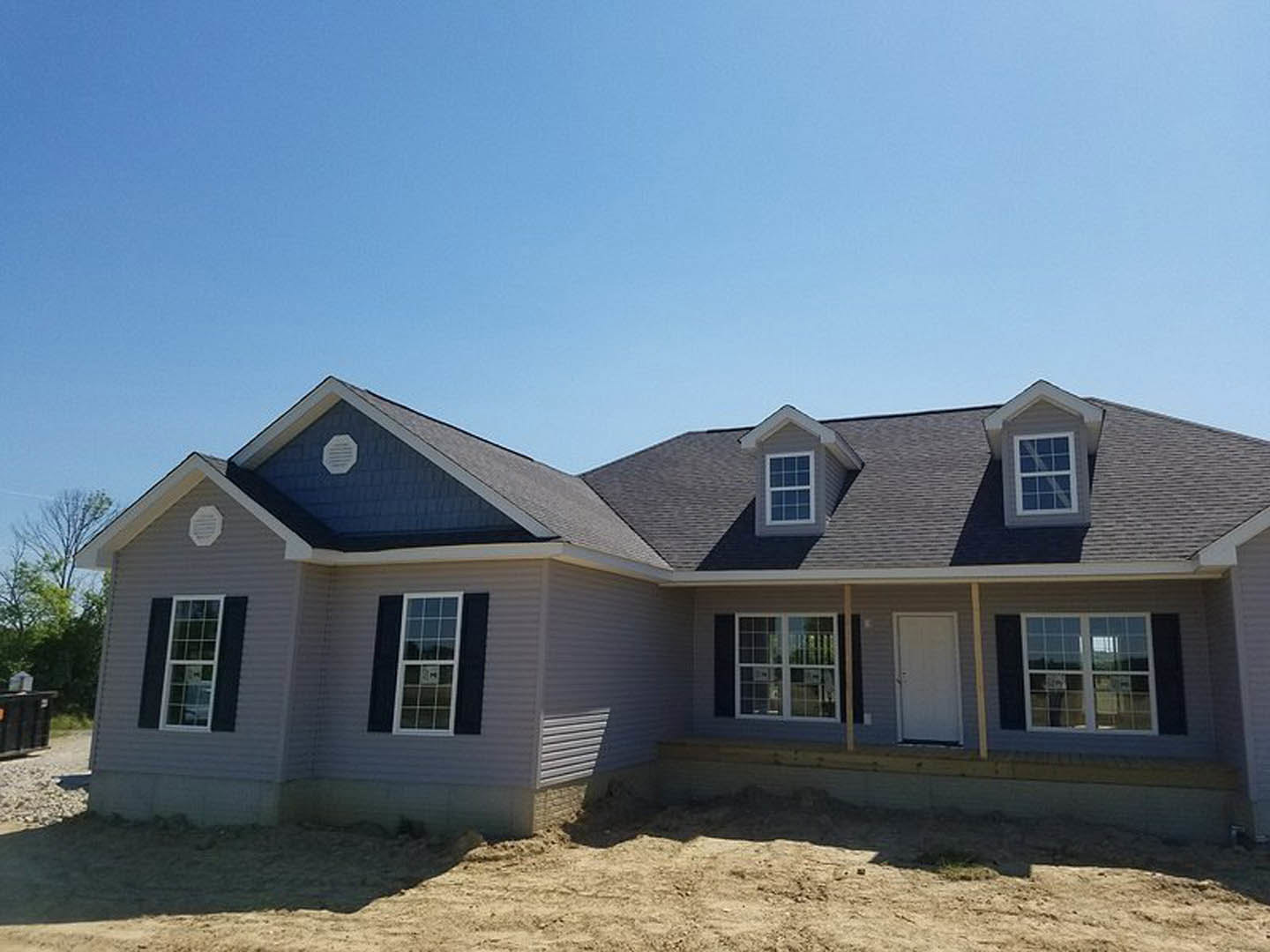 Grey house under construction with white framed windows, white door, dirt yard, and blue sky overhead