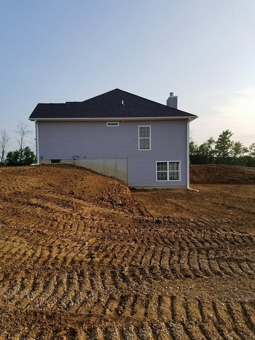 Light-colored cottage with gabled roof, large front window displaying a sign, surrounded by green trees and plants, set beside a dirt hill under open sky.
