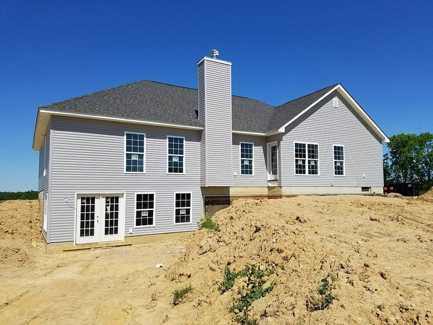 White custom home under construction with double glass doors, gray siding, and a dirt pile in front; young plants and a leafy tree visible near the foundation.