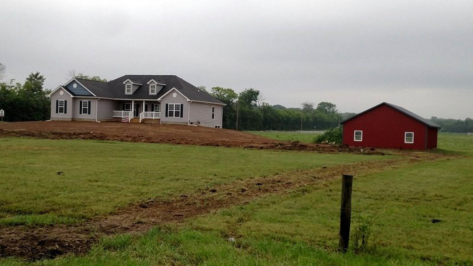 Two-story farmhouse with white railing and white-trimmed windows, red barn siding, surrounded by grassy field and scattered trees under a partly cloudy sky