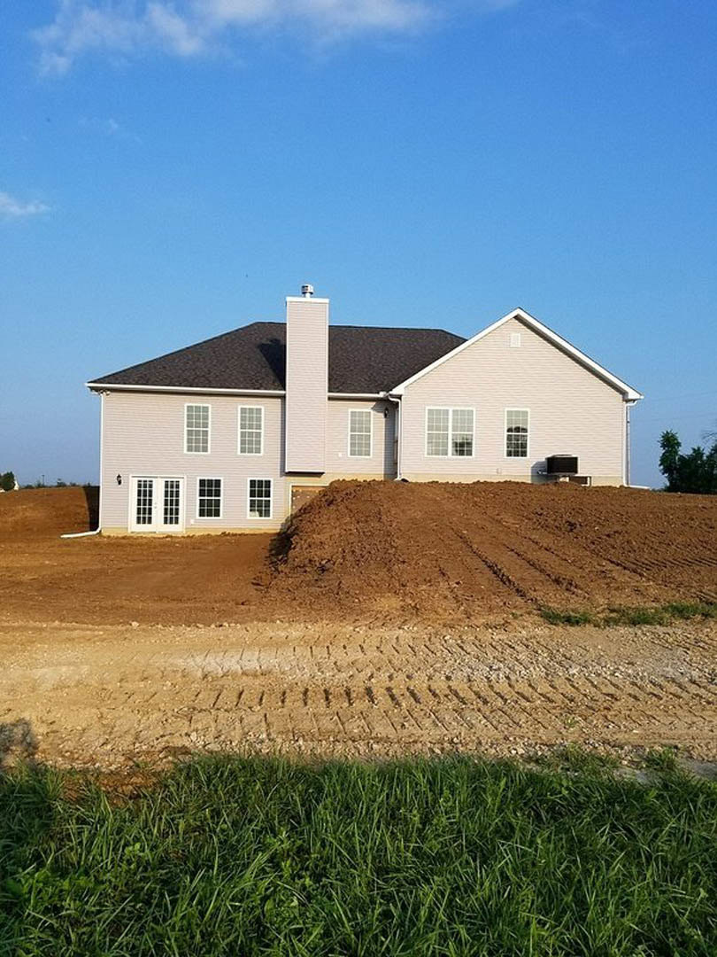 Partially built house with exposed framing, surrounded by dirt and patches of grass, chimney visible, cloudy sky overhead
