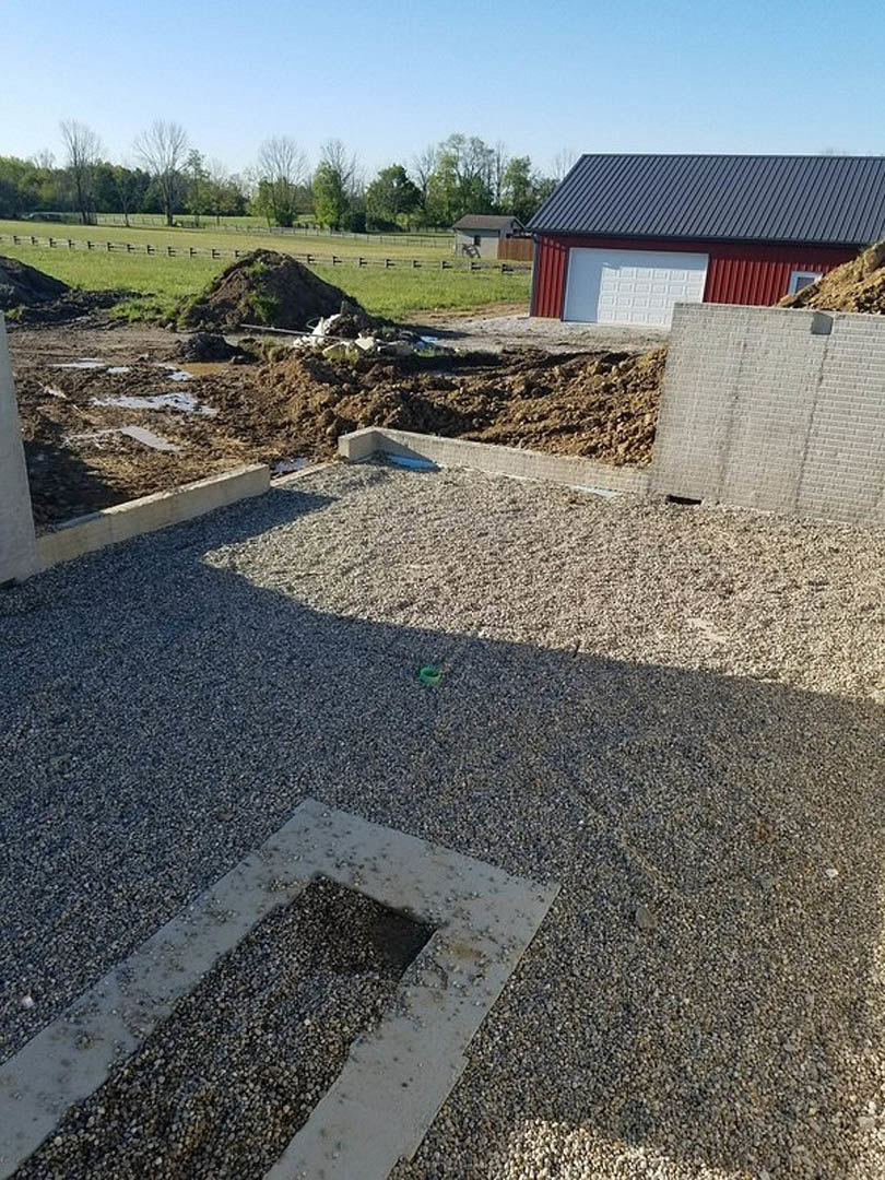 Two-story custom home under construction with exposed brick walls, white siding, black window frames, gravel driveway, and dirt lot surrounded by trees.