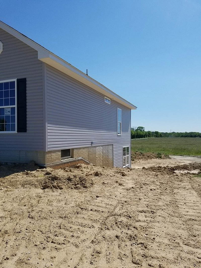 Two-story home with white siding, large windows, dirt pile in front yard, patches of grass, and trees in the background under a clear blue sky