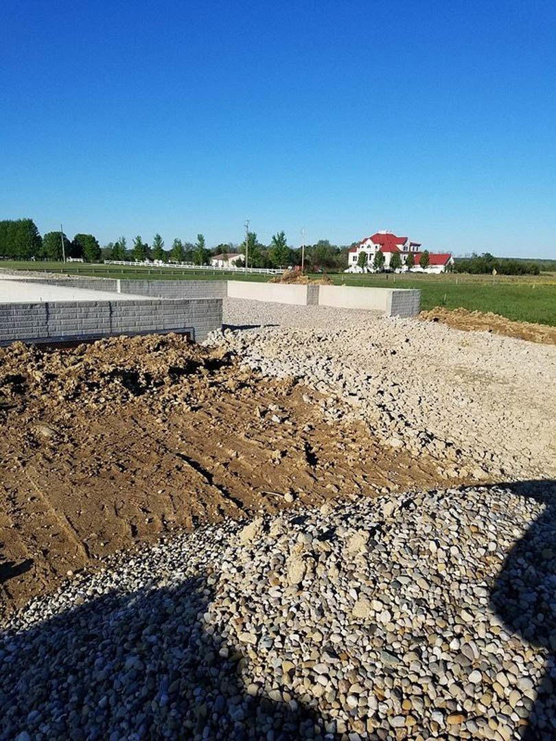 Large white house with red roof sits in open field with dirt and gravel foreground, pile of rocks, cracked wall, and blue sky overhead
