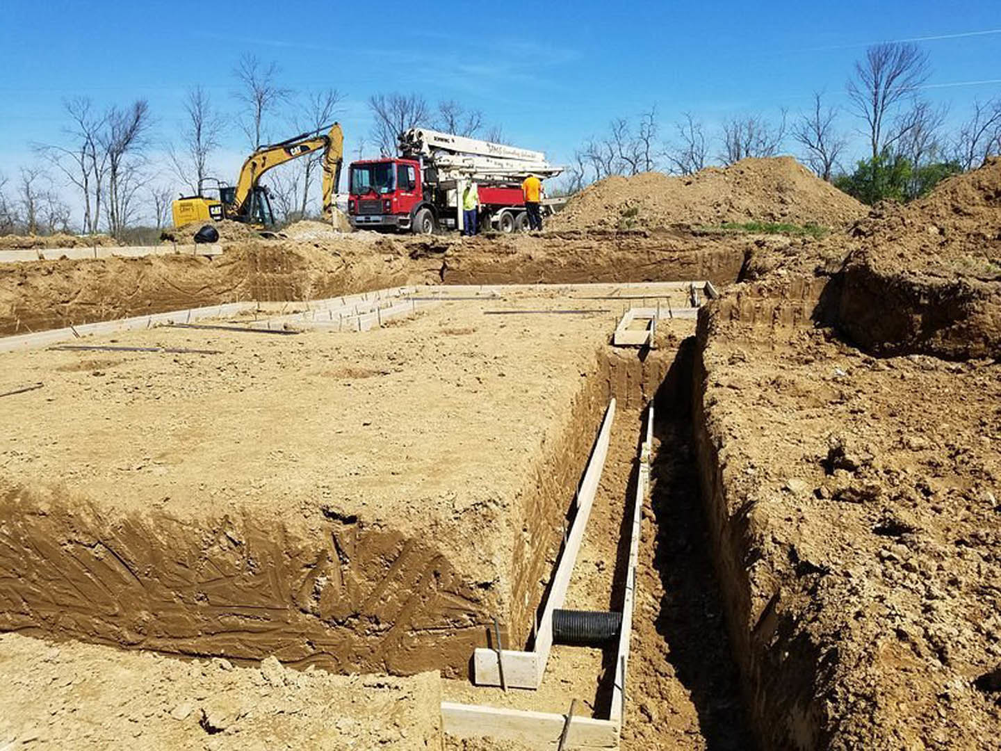 Construction site with exposed soil, yellow excavator, two trucks—one with crane attachment—man standing beside vehicle, trench with visible pipe, tree line in background