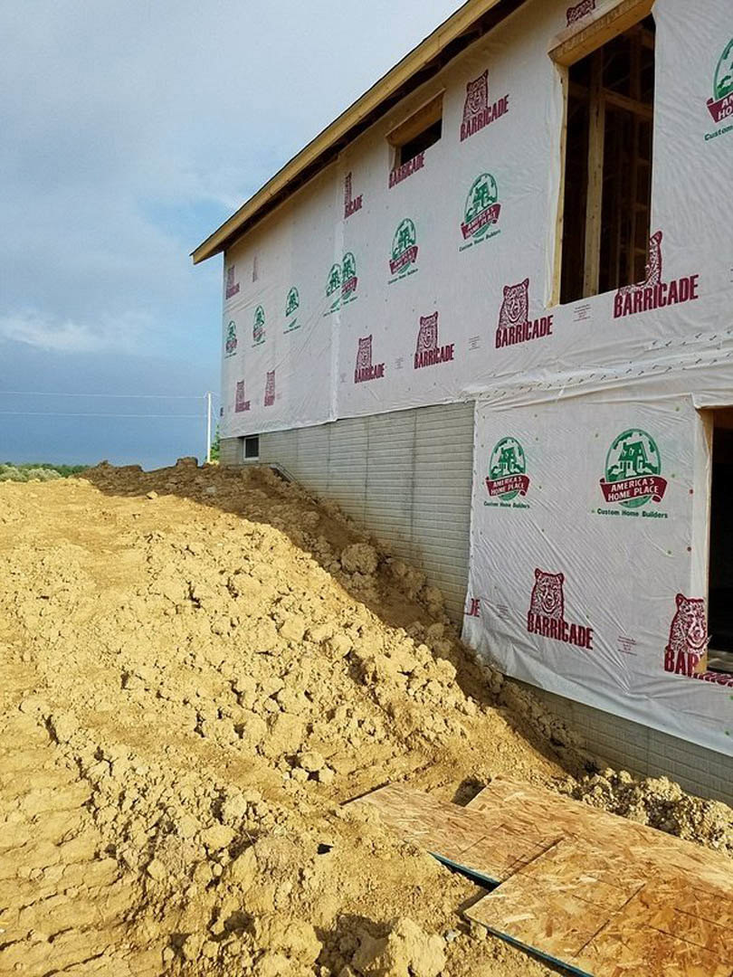 Wood-framed custom home under construction with exposed plywood walls, window openings, and a large pile of dirt in the foreground beneath a partly cloudy blue sky