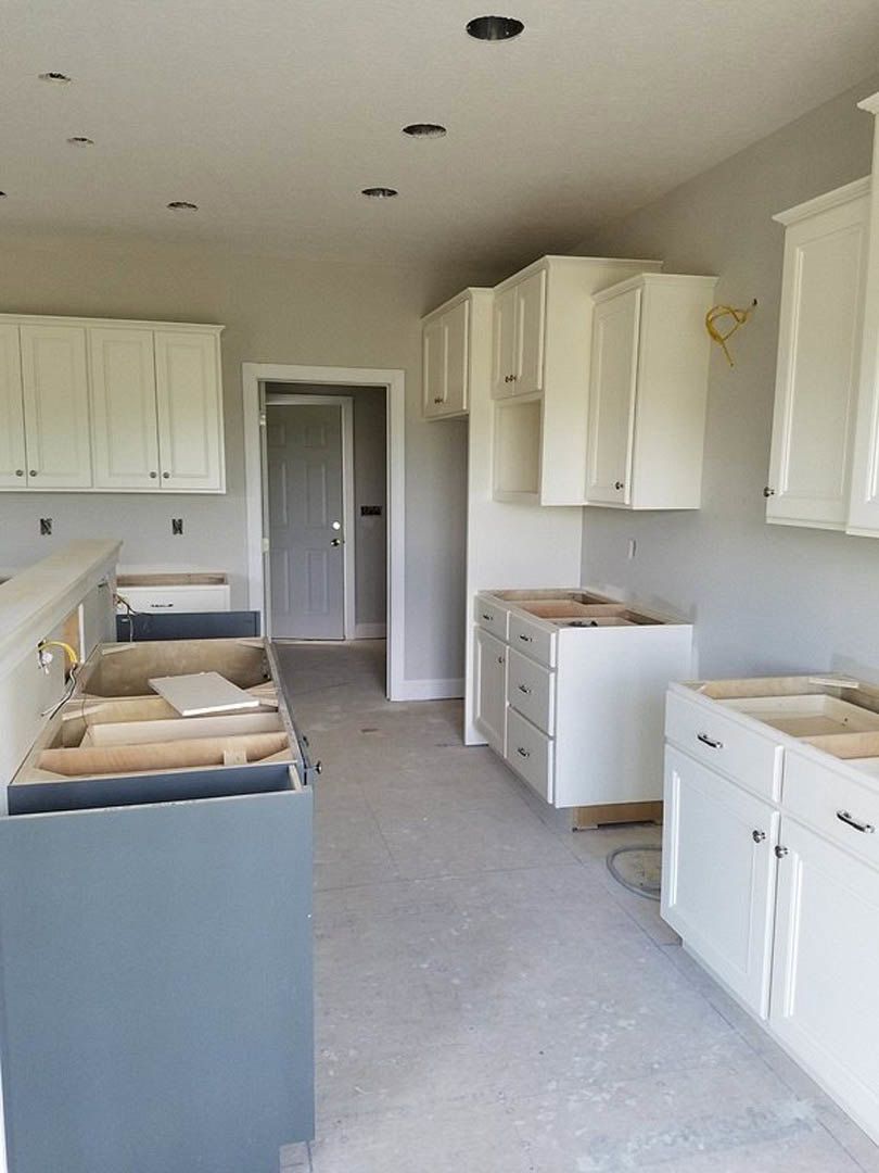 White kitchen with shaker-style cabinets and drawers, silver knobs, white door with round knob, yellow wire along white wall, wood piece in machine, light-colored flooring.