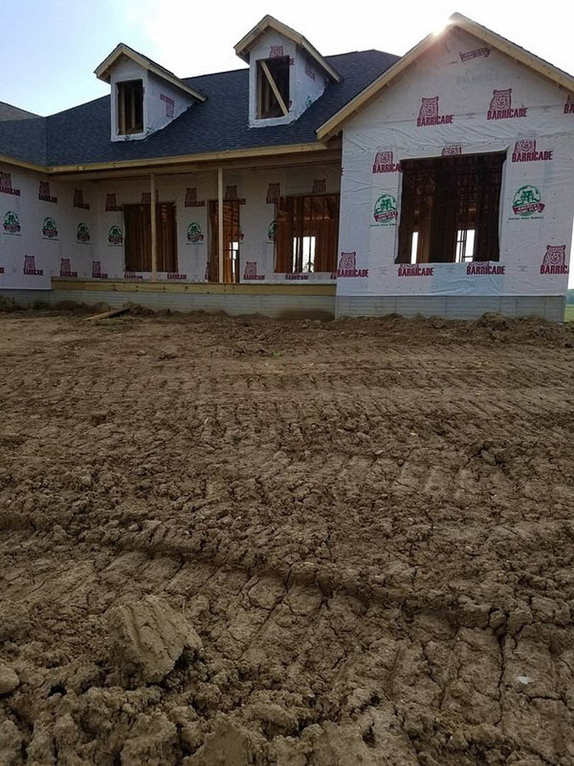 Partially built house with exposed framing, several installed windows, dirt ground with tire tracks, white curtain visible through window, construction materials scattered outside
