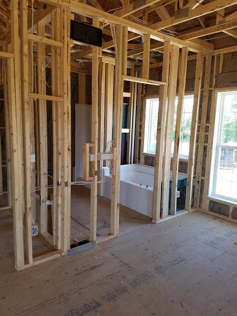Bathroom under construction featuring exposed wood framing, white-framed window, built-in bathtub, wall-mounted black screen, and unfinished flooring