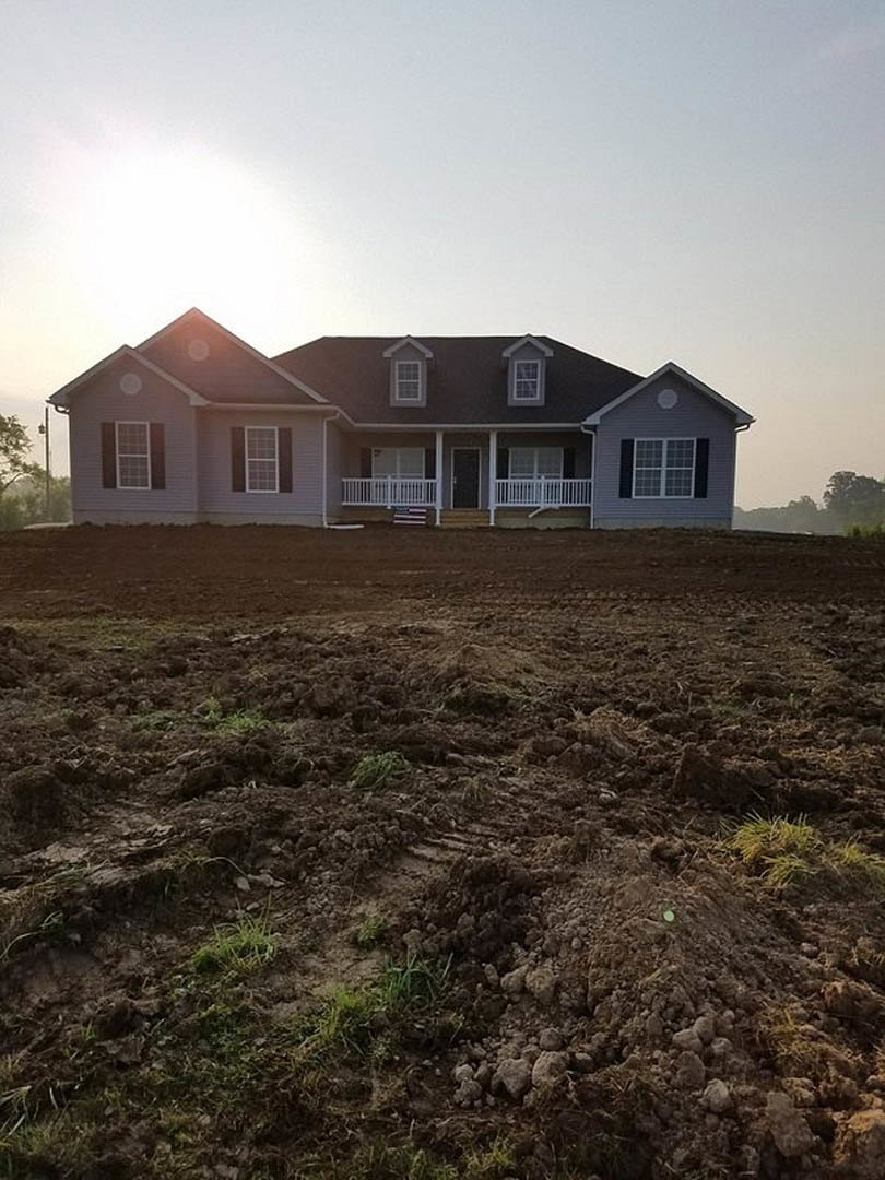 Two-story house with gray roof, white porch railing, and multiple square windows, surrounded by a large dirt field with visible tractor tire tracks, under a clear sky.