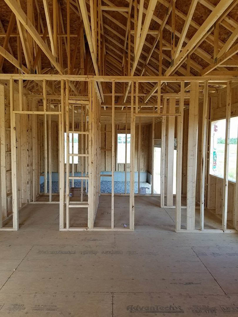 Exposed wooden beams and framing with unfinished floor, large window featuring a green construction sign, natural daylight illuminating interior structure