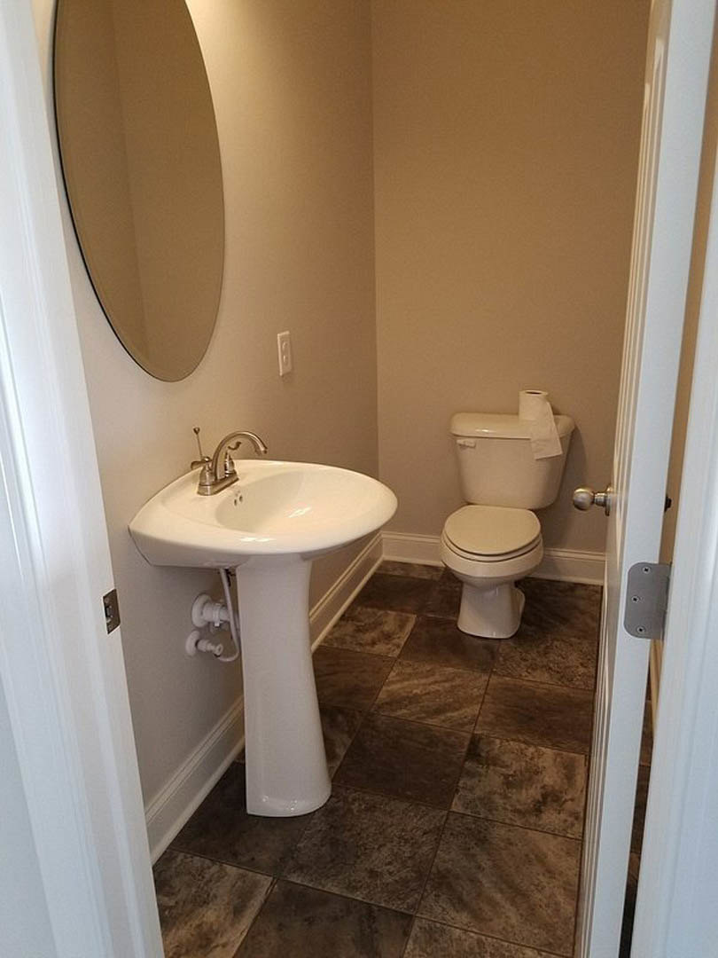 Bathroom with white tile walls, round mirror above a wall-mounted sink with exposed plumbing, chrome faucet, and toilet with lid up positioned beside the sink.