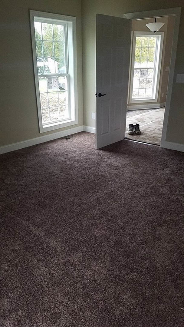 White door with black handle open to carpeted room, white-framed window with sign visible outside, light-colored walls, wood laminate flooring in foreground.