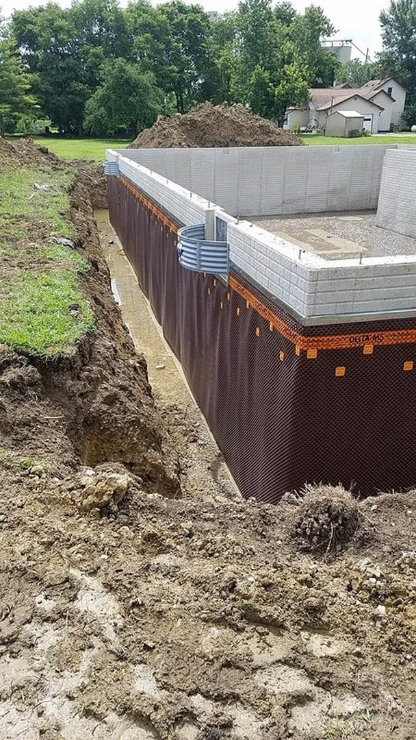 Trench in soil beside a foundation wall covered with black and brown fabric, outdoor setting with grass, trees, and sky visible