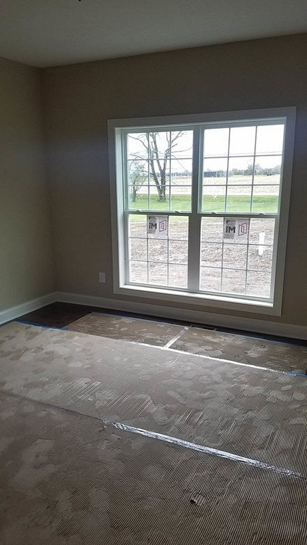 Carpeted room with a large window overlooking a field and trees, white baseboard trim along plaster walls, natural daylight illuminating black flooring with a white accent line