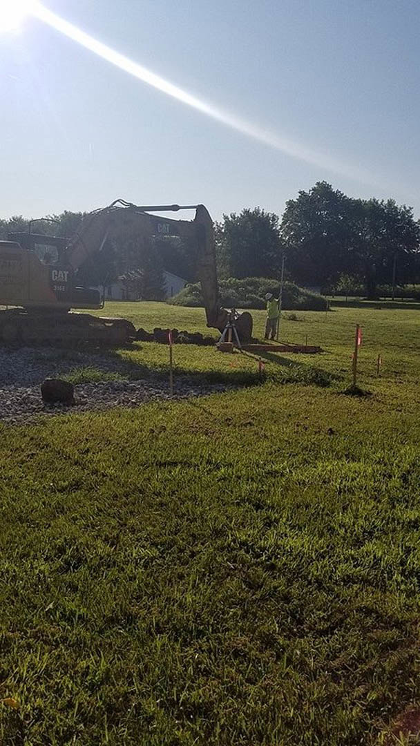 Large yellow excavator digging earth on grassy residential construction site, surrounded by open field, trees, and cloudy sky