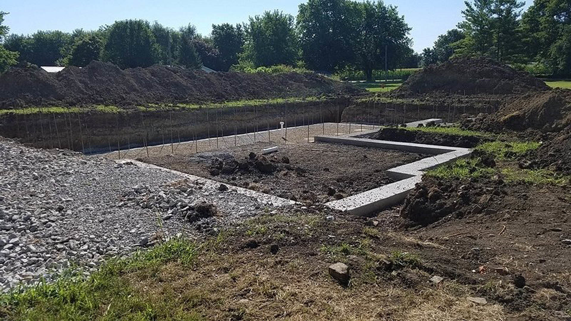 Concrete house foundation surrounded by soil, rocks, and sparse groundcover plants with trees and blue sky in the background
