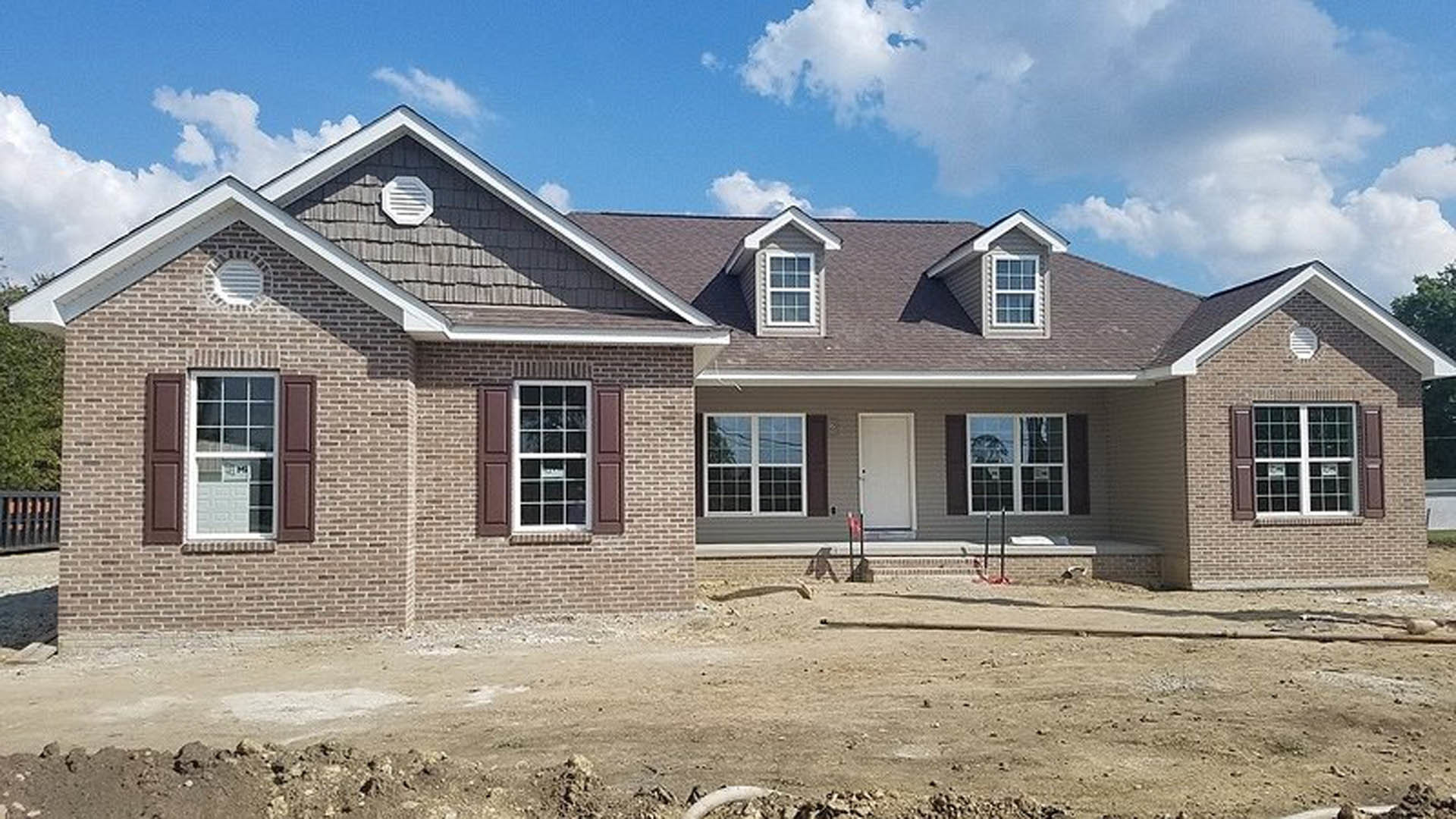 Wood-framed house under construction with exposed siding, unfinished roof, dirt foreground, and blue sky with scattered clouds