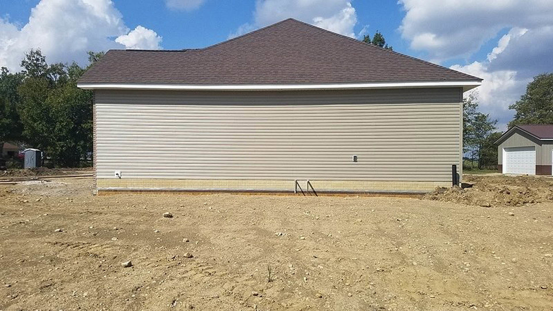 White garage door on a newly constructed home, dirt yard in foreground, portable toilet visible to the side, cloudy sky overhead