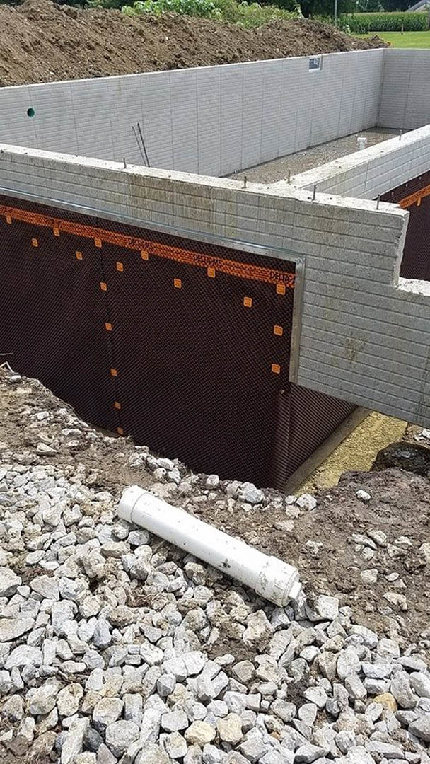 Concrete wall with orange and black panels, white pipe resting on rocky ground at residential construction site