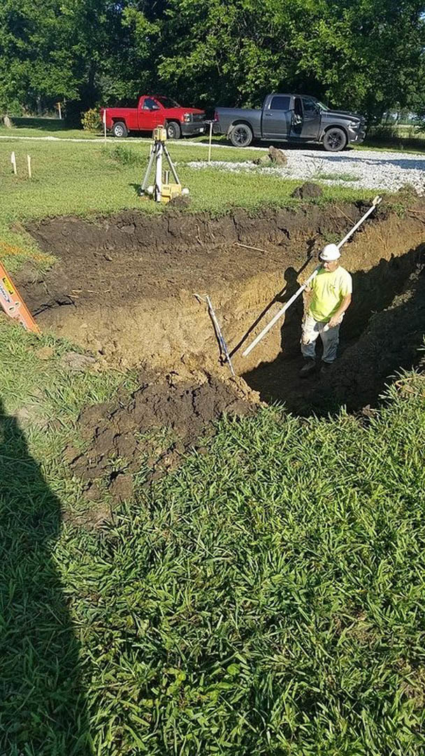 Man standing in a soil excavation on grassy lawn, grey truck with open door and red truck parked nearby, gas cylinder and person in yellow shirt visible, trees and plants in