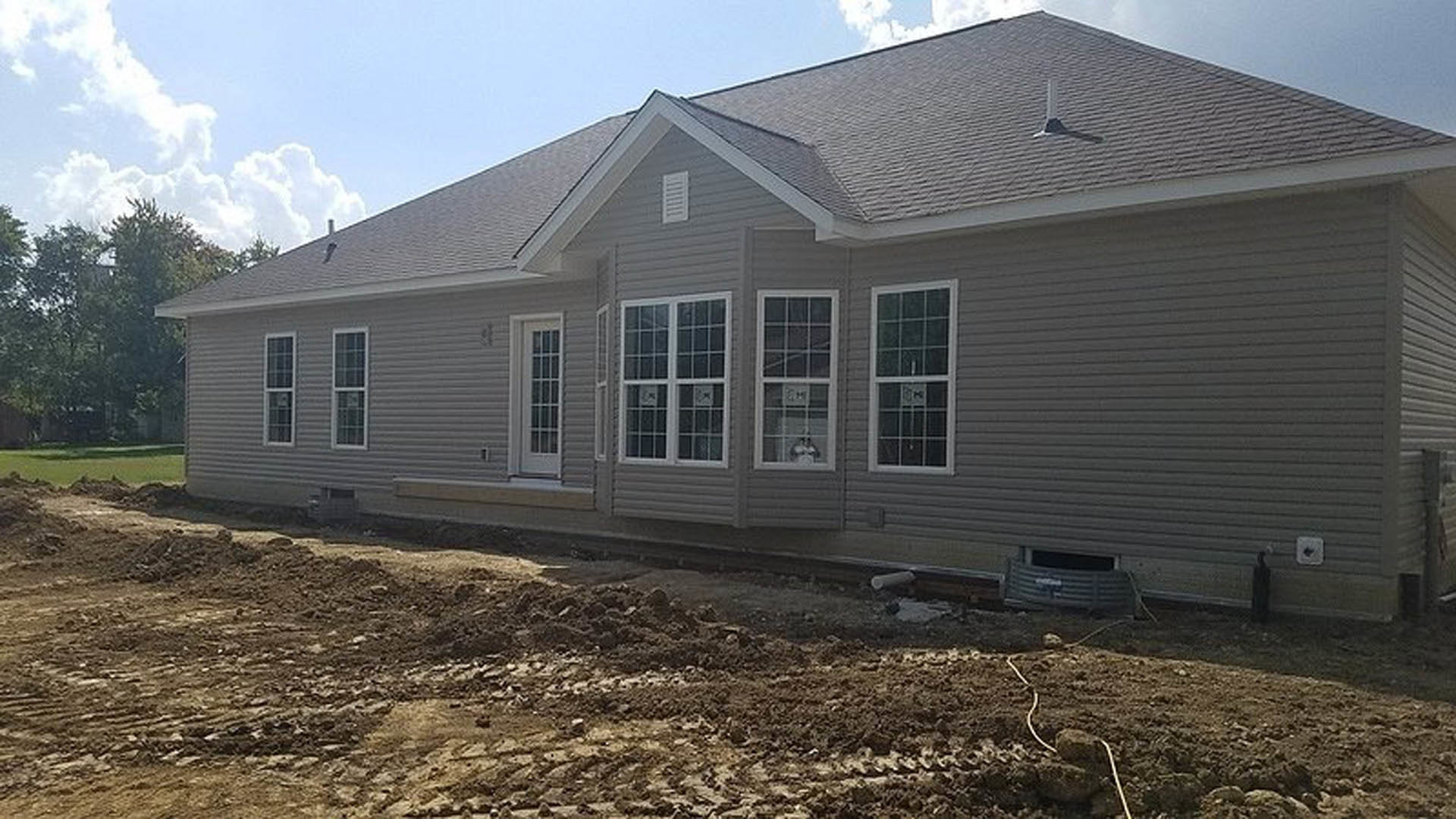 Gray house under construction with multiple pane windows, dirt ground in foreground, white window trim, cloudy sky above