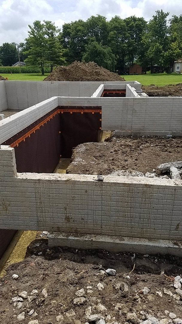 Concrete foundation with exposed excavation surrounded by grass, trees, and outdoor construction materials under a clear sky