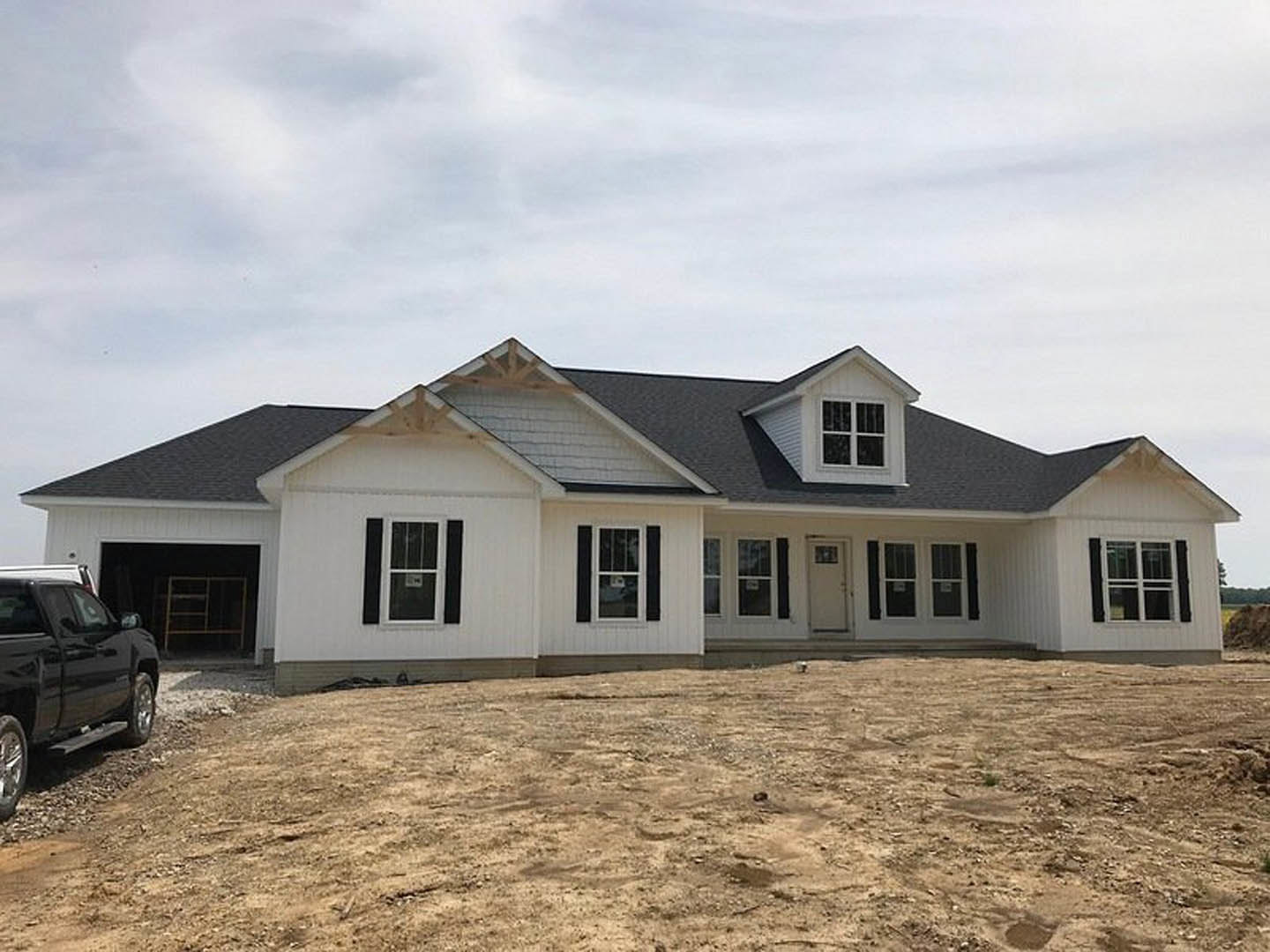 White house under construction with black shutters, dirt foreground, blue sky, black truck parked nearby, white door with black frame, white-framed window