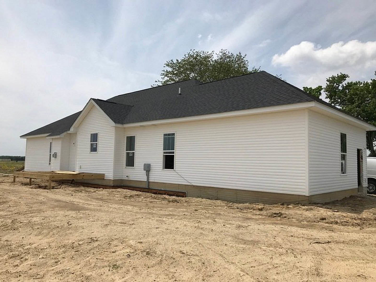 Partially built house with white-framed window, exposed roof, dirt yard, and surrounding trees
