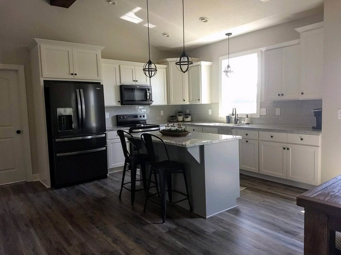 White kitchen cabinets, black double-door refrigerator, black microwave, wood table, two black chairs at kitchen counter, light flooring
