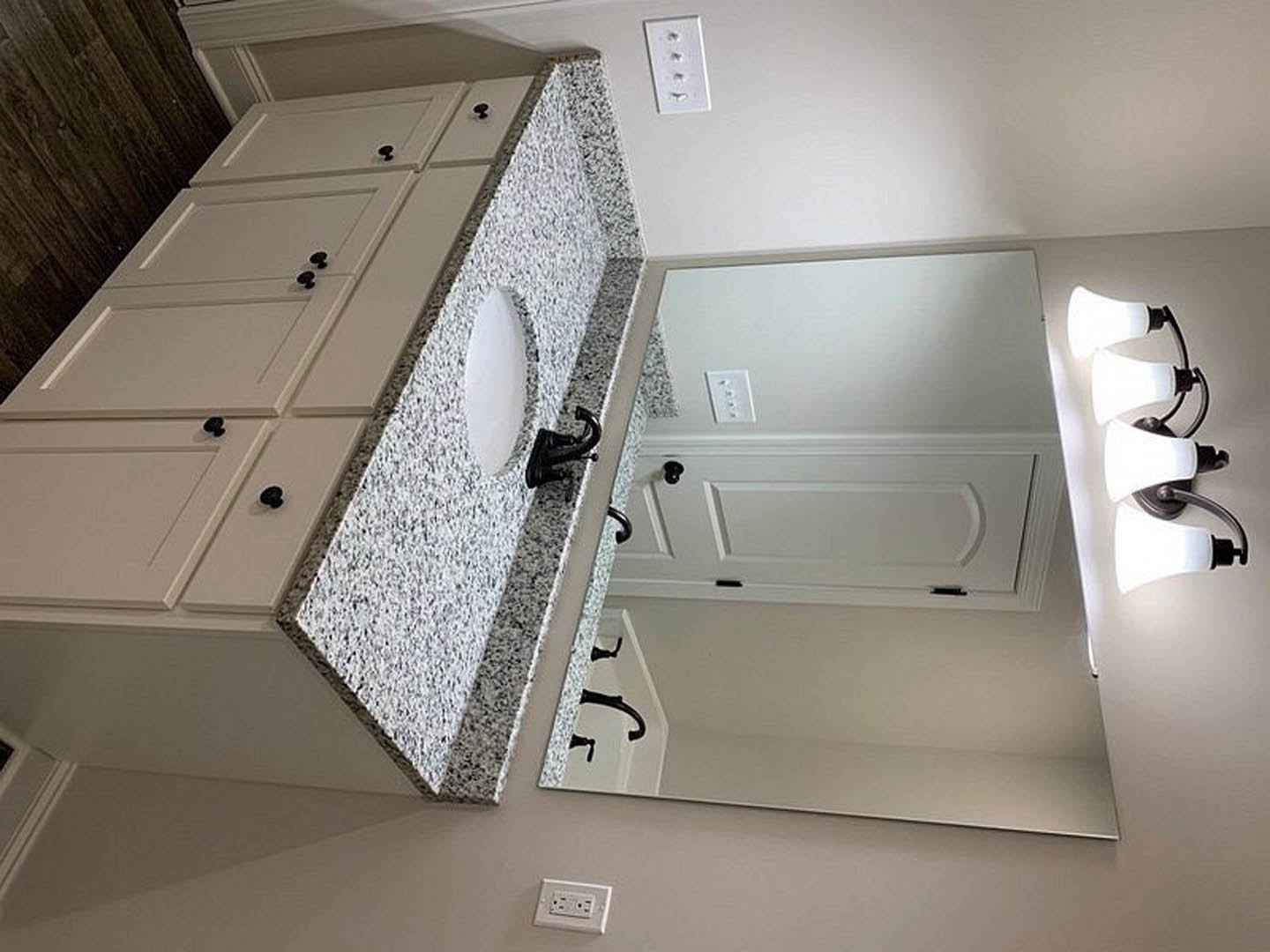 Modern bathroom with white sink, rectangular mirror, black and white light fixture above, white wall outlet with four switches, and white door featuring a black knob