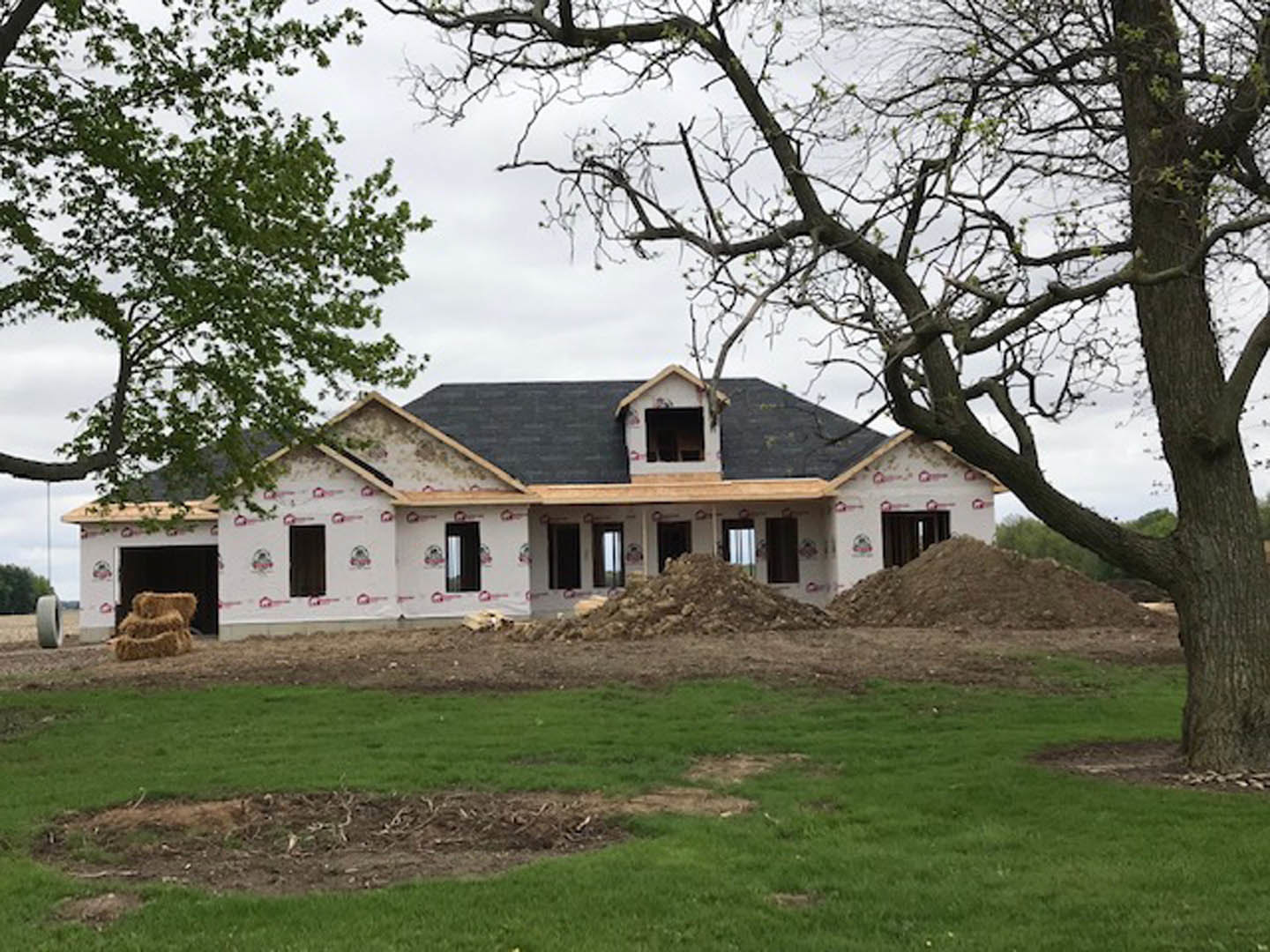 Wood-framed house under construction surrounded by dirt, grass, and mature trees under a clear sky