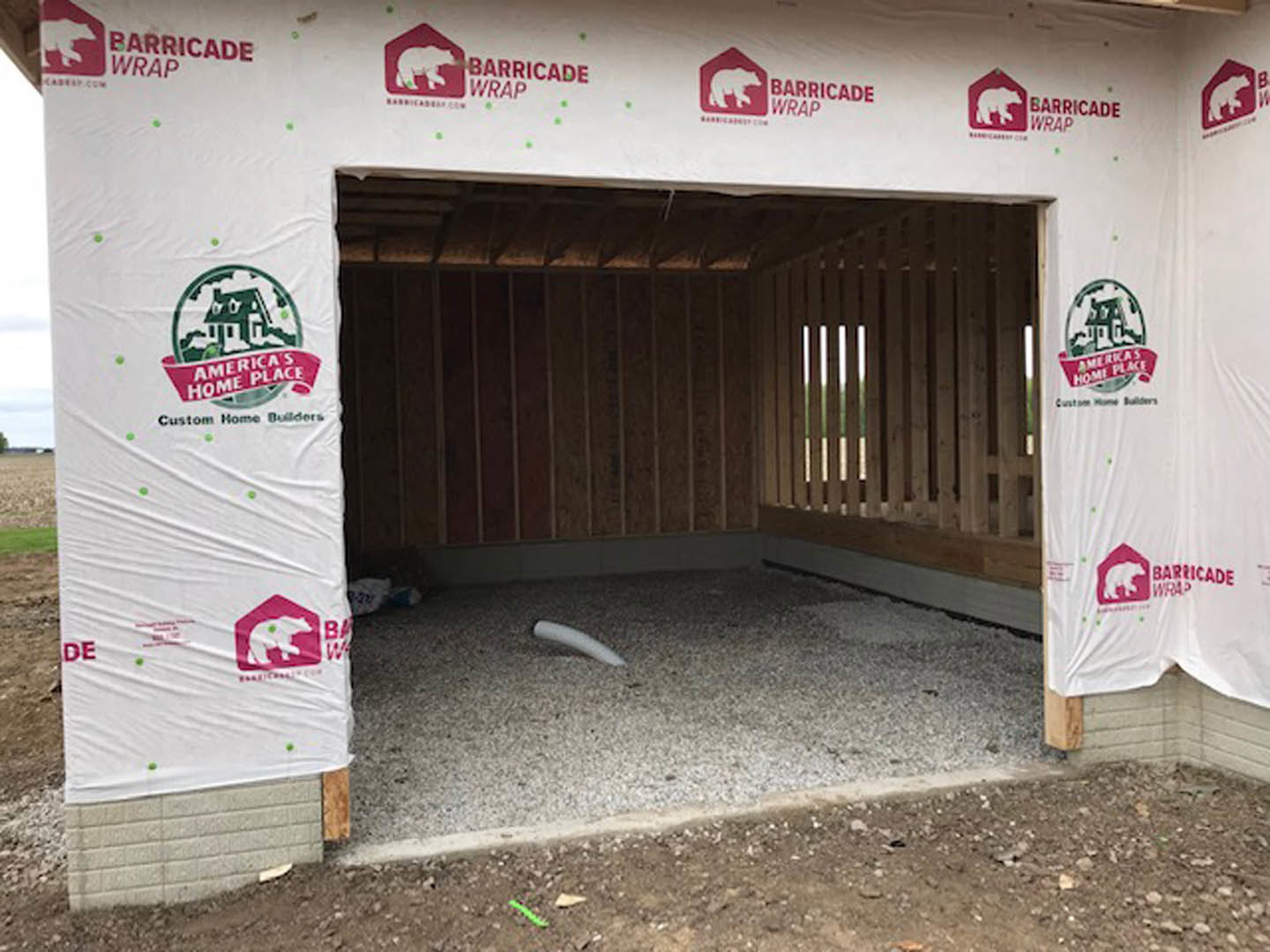 Partially constructed residential building wrapped in white protective sheeting, exposed ground and construction materials visible near entry door.