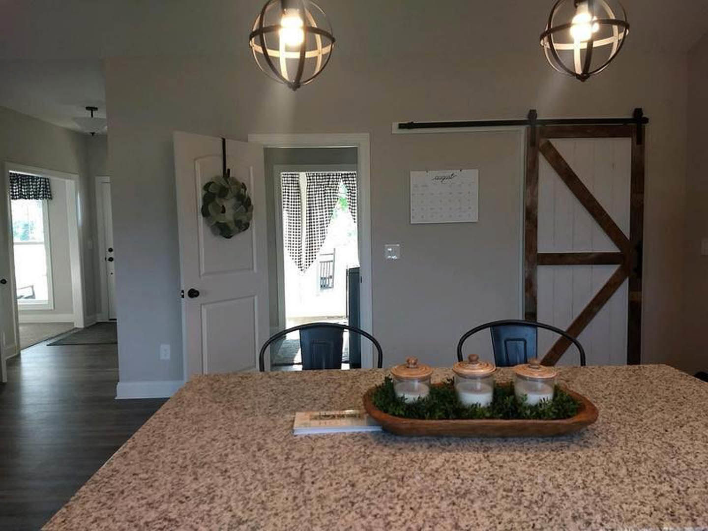 Dining room with wooden table set with candles, glass jar, and decorative bowl; white door framed by brown wooden beams, wall calendar, and upholstered chair visible in background.