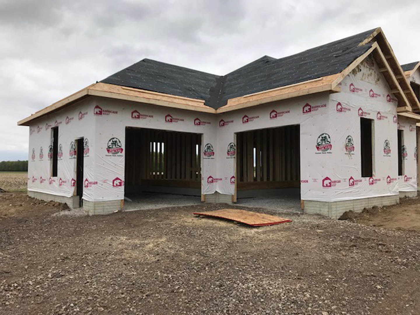 Partially built house with completed shingle roof, exposed wooden framing, and construction materials scattered on dirt ground; windows installed, cloudy sky overhead.