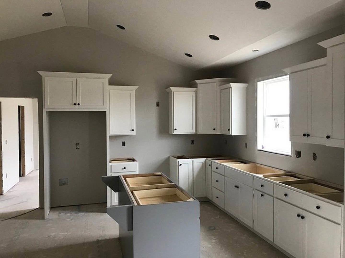 White kitchen cabinets with black knobs, light gray countertop, stainless steel sink, close-up of a drawer with interior shelf, window above sink, wall-mounted light switch.