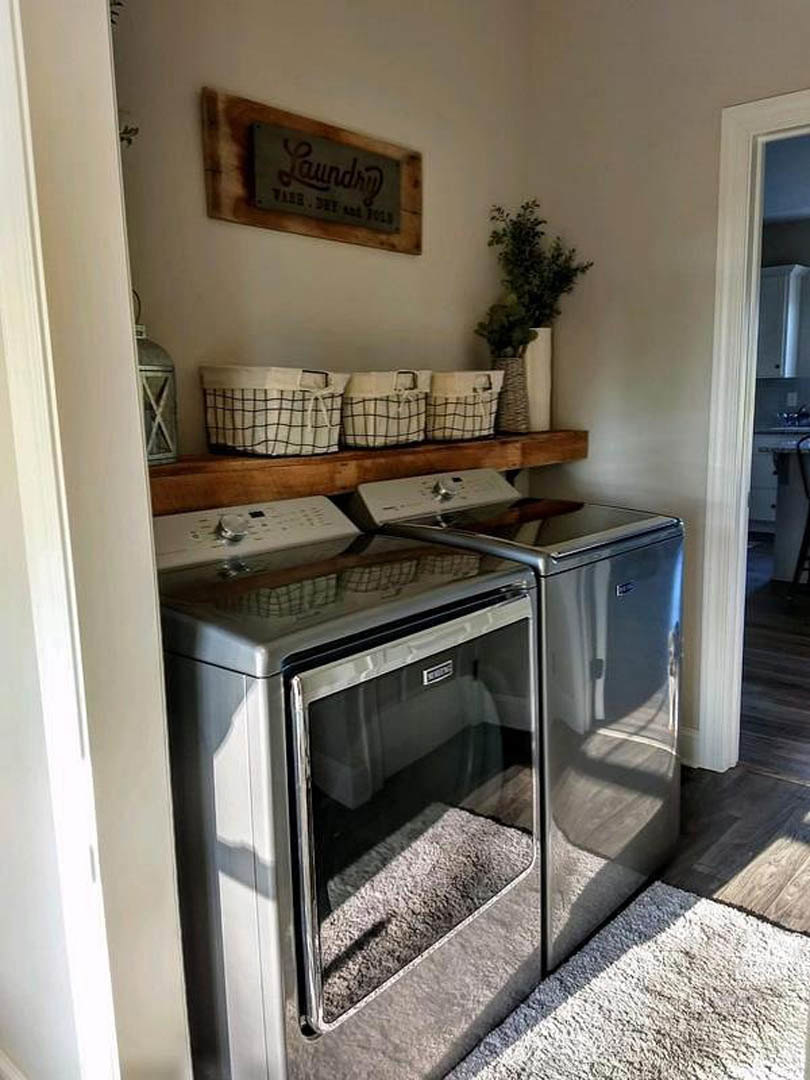 Front-loading washer and dryer set against a white wall with a decorative sign, potted plant in a glass vase on the countertop, black and white woven laundry baskets, and a