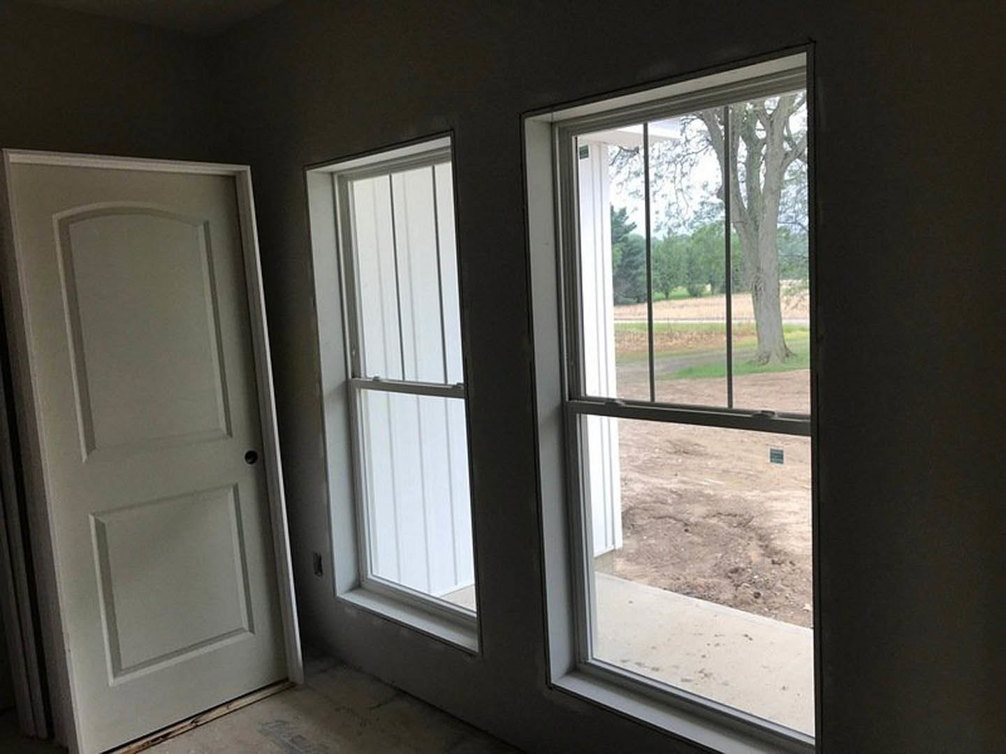 Bright room with two large windows, white walls, light wood flooring, white door with black handle, view of dirt field and trees outside