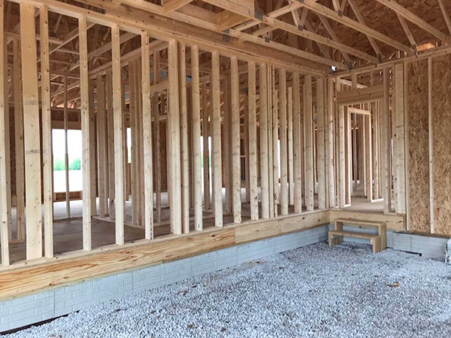 Unfinished interior with exposed wooden beams, gravel flooring, and a partially constructed wooden bench with shelf