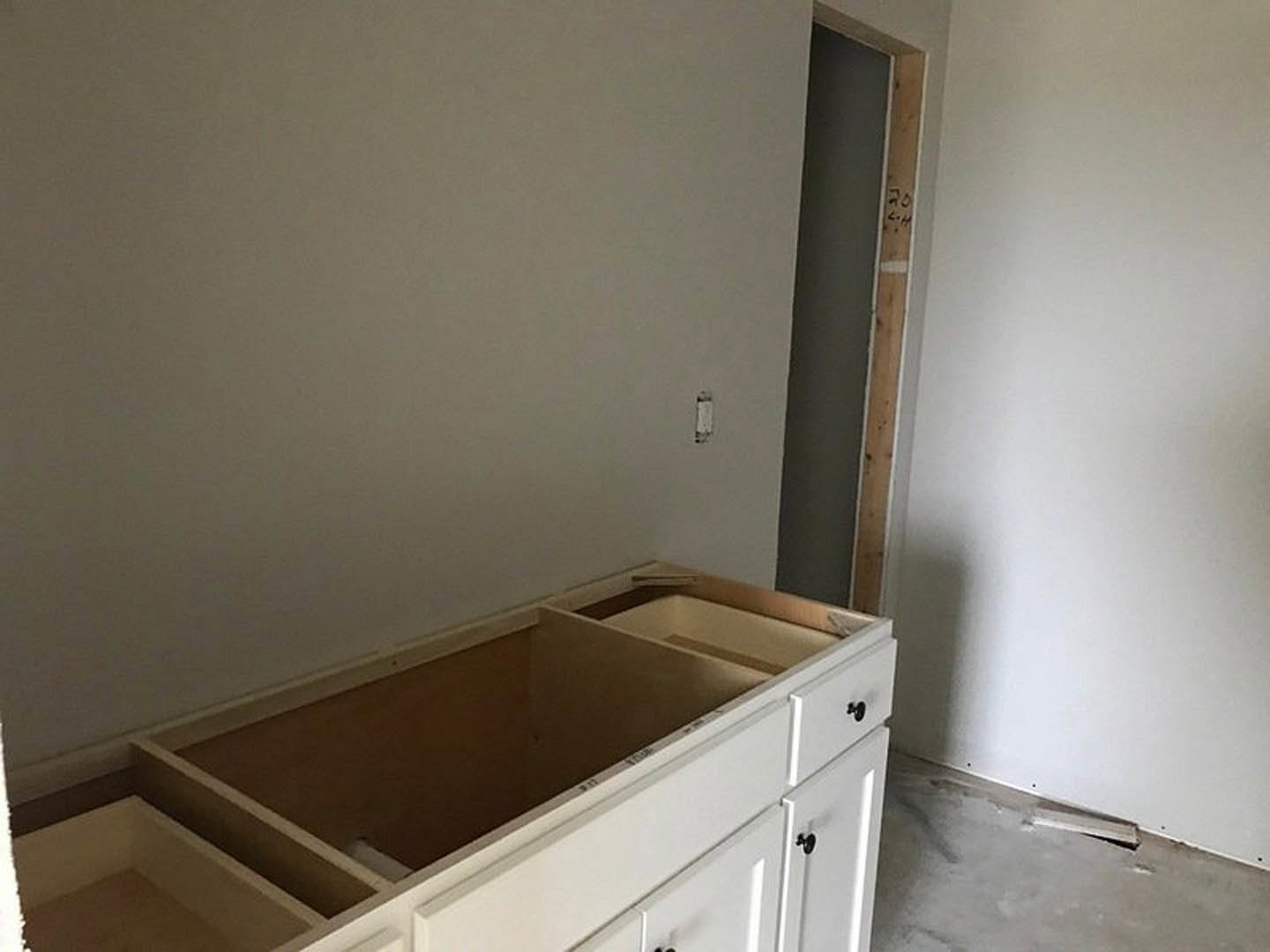 White kitchen cabinetry with shaker-style drawers, white walls, light wood flooring, and exposed wooden ceiling beam in a corner of the room.