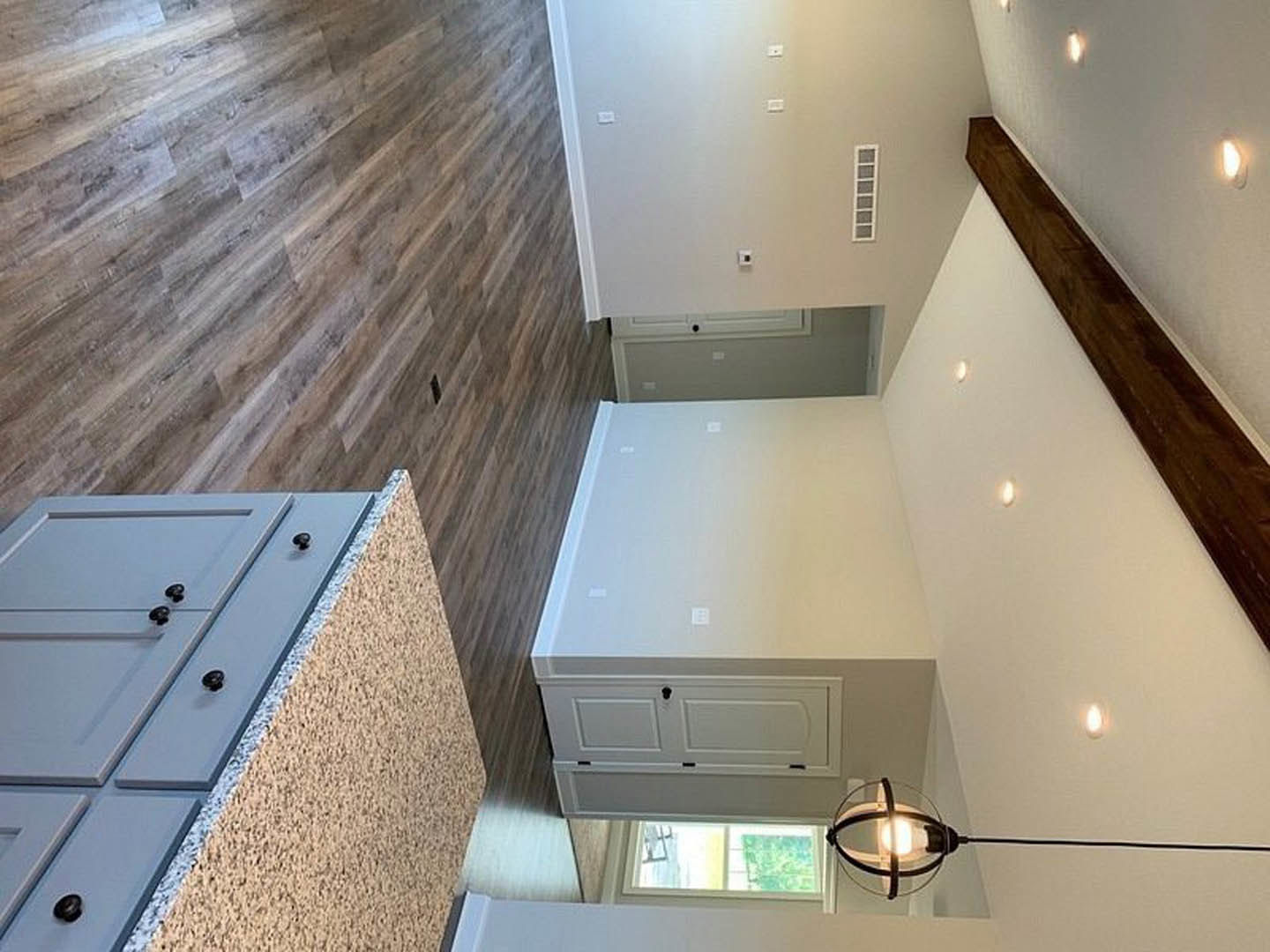 Kitchen with warm wood flooring, white cabinetry featuring a keyhole, round light fixture, smooth white walls, and a partially visible window.
