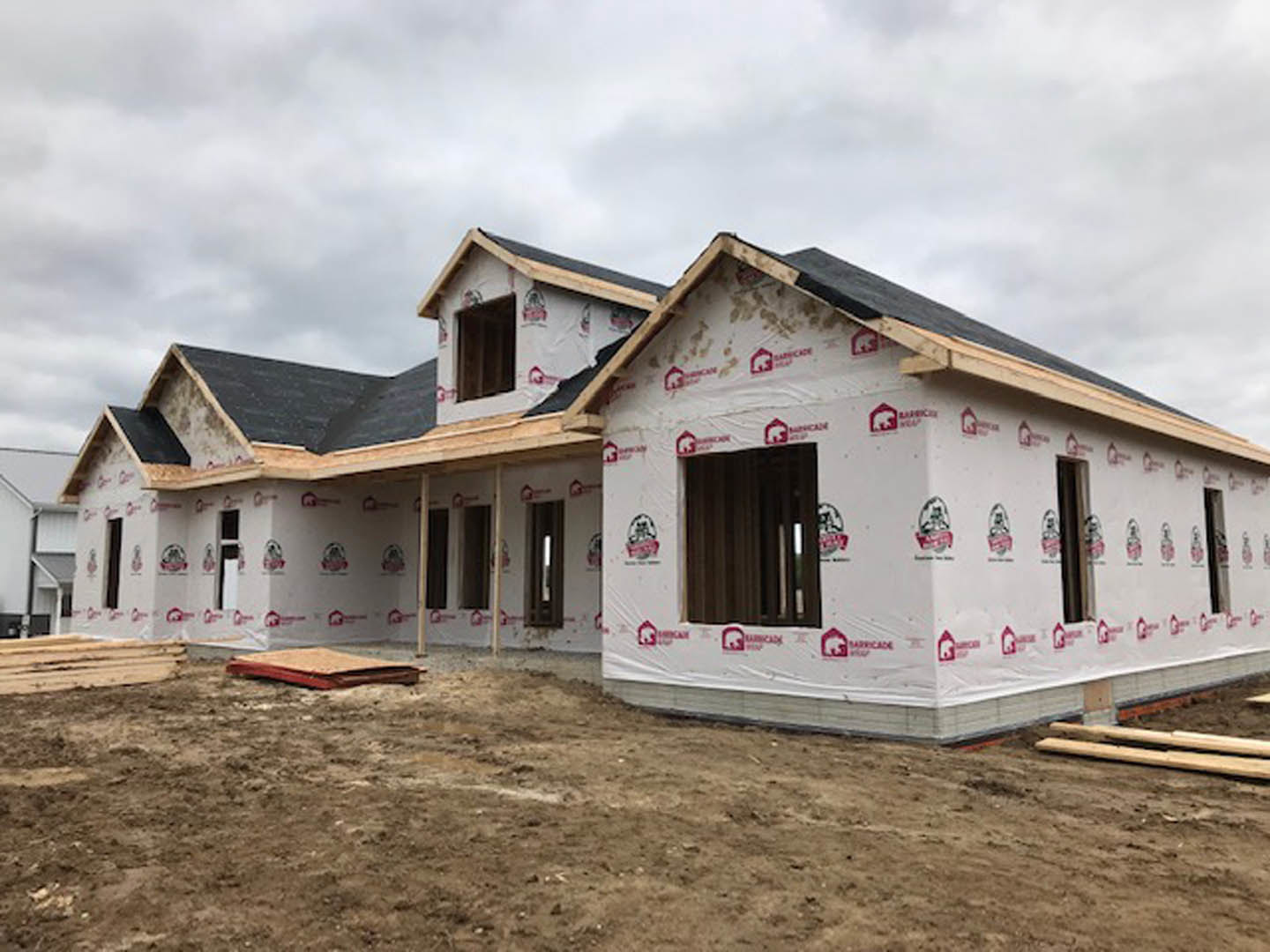 Partially built house with exposed framing, several installed windows, white construction materials scattered on dirt ground, cloudy sky overhead