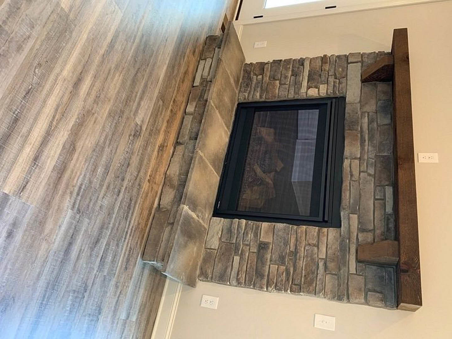 Stone fireplace with wooden mantel, wood flooring, plaster walls, large window, and white electrical outlet with black button