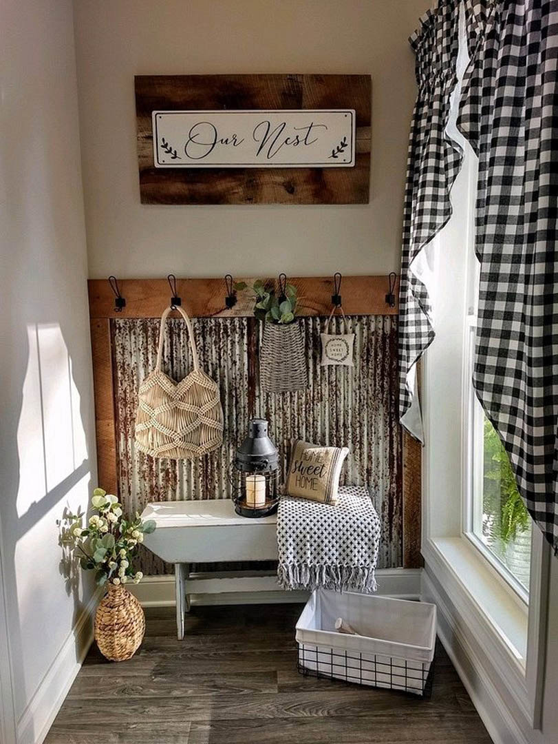 Entryway with a wooden bench, woven basket holding a white cloth, lantern-style lamp, and a vase of fresh flowers against a light-colored wall with sheer curtains.