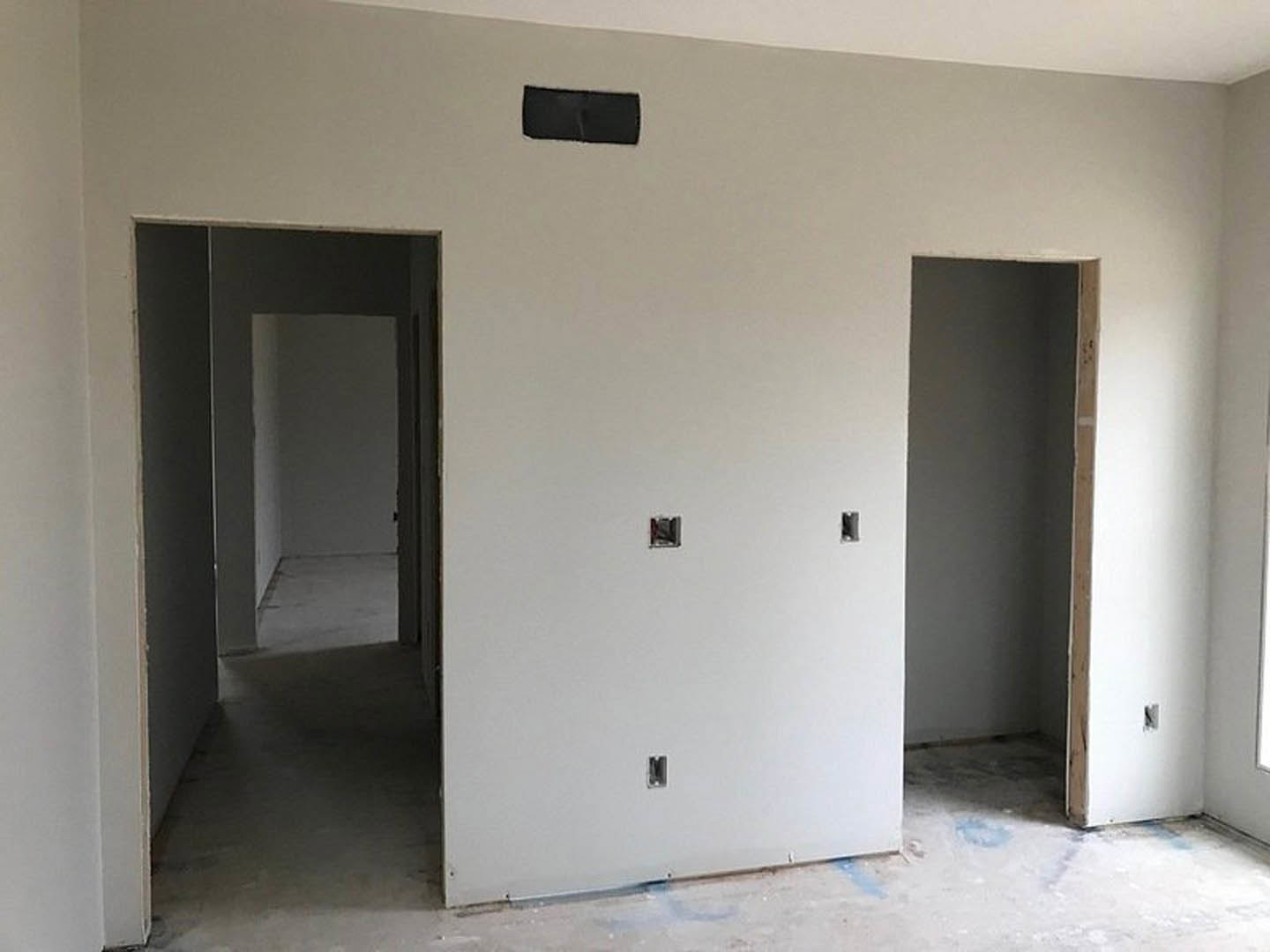 White-walled room with visible holes, open doorway leading to hallway, wooden door frame, black and red electrical outlet, light-colored flooring, and ceiling.