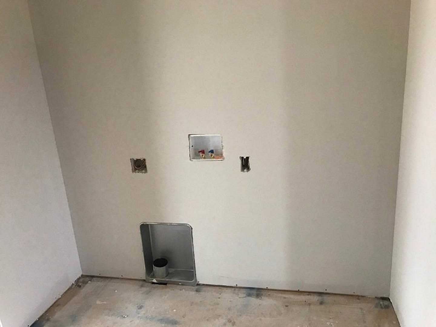 White plaster bathroom wall with a rectangular hole, exposed metal pipe in built-in metal shelf, silver trash can with black lid, close-up of floor vent, concrete flooring, small