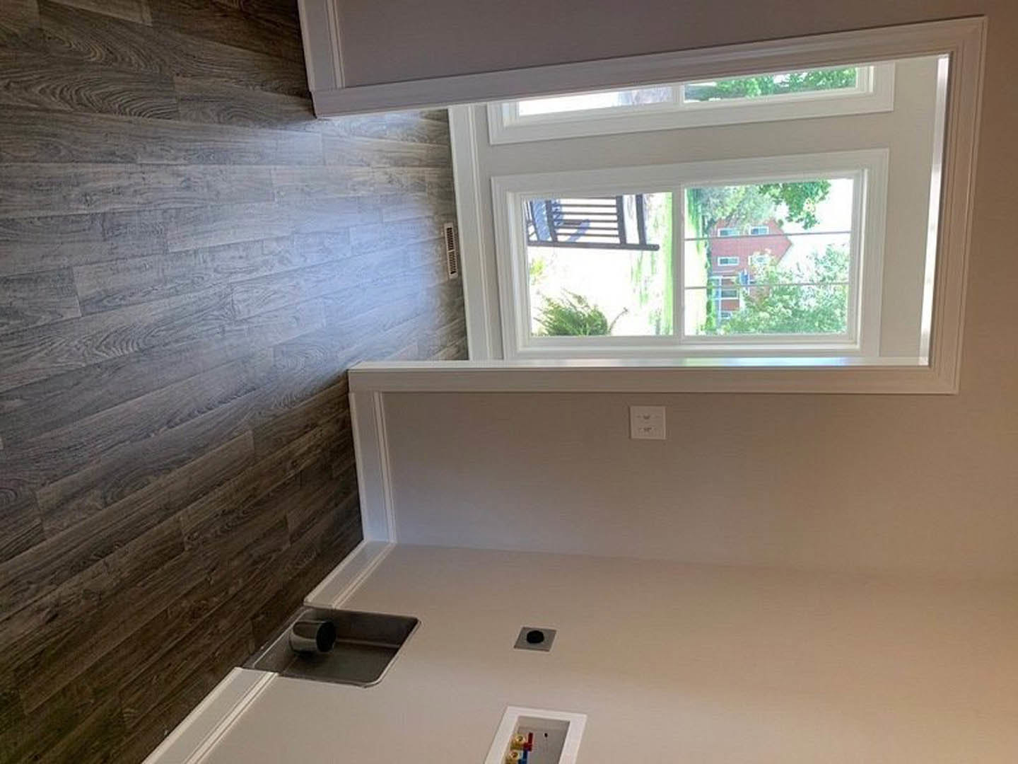 Sunlit room with wood flooring, plaster walls, large window overlooking a house and trees, white wall switch, wood accent wall with white railing, bench and cup in foreground.