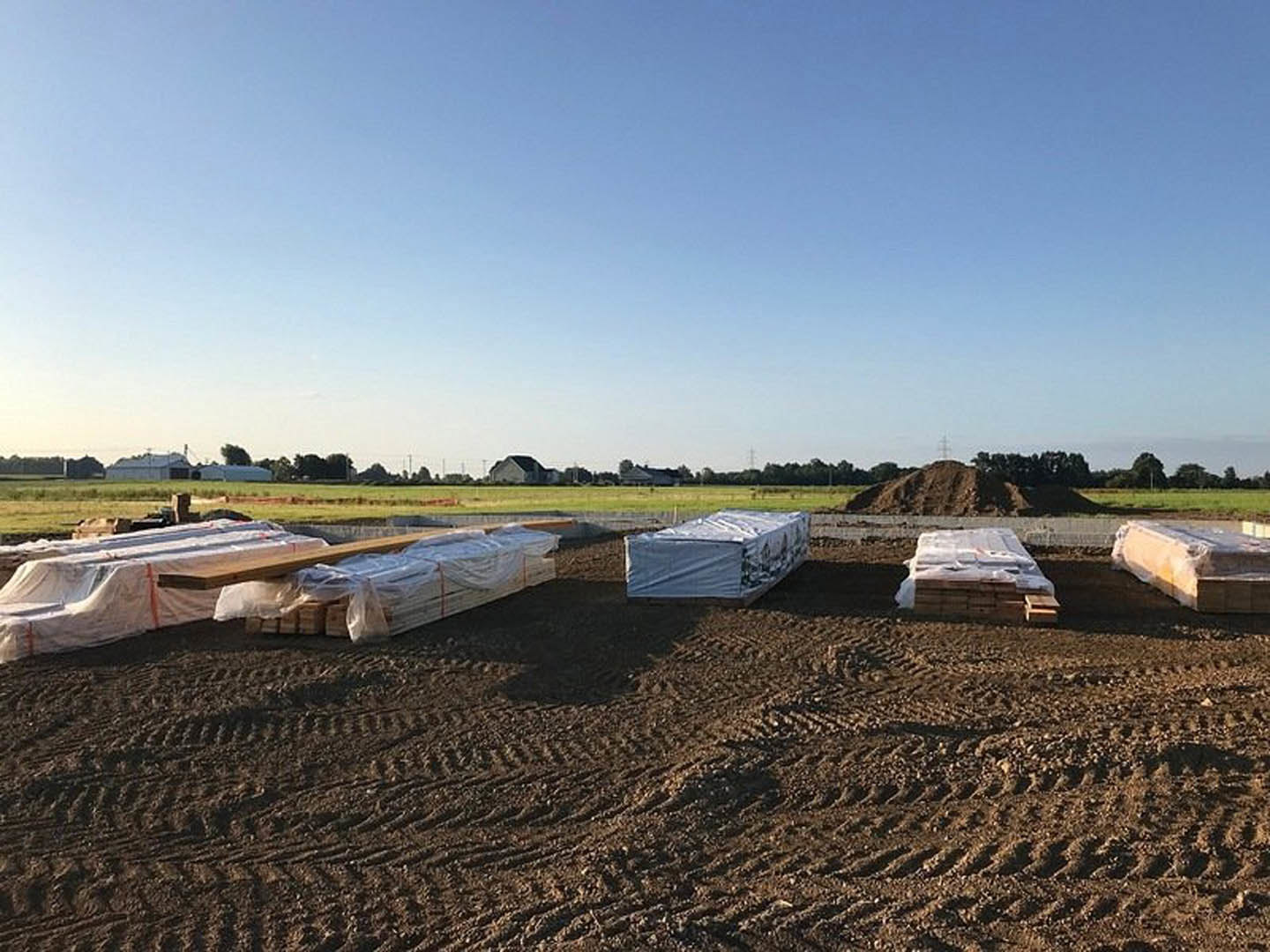 Stacked wooden pallets and boxes on a dirt field, large rectangular object covered with a tarp, scattered concrete blocks, clear blue sky with clouds overhead