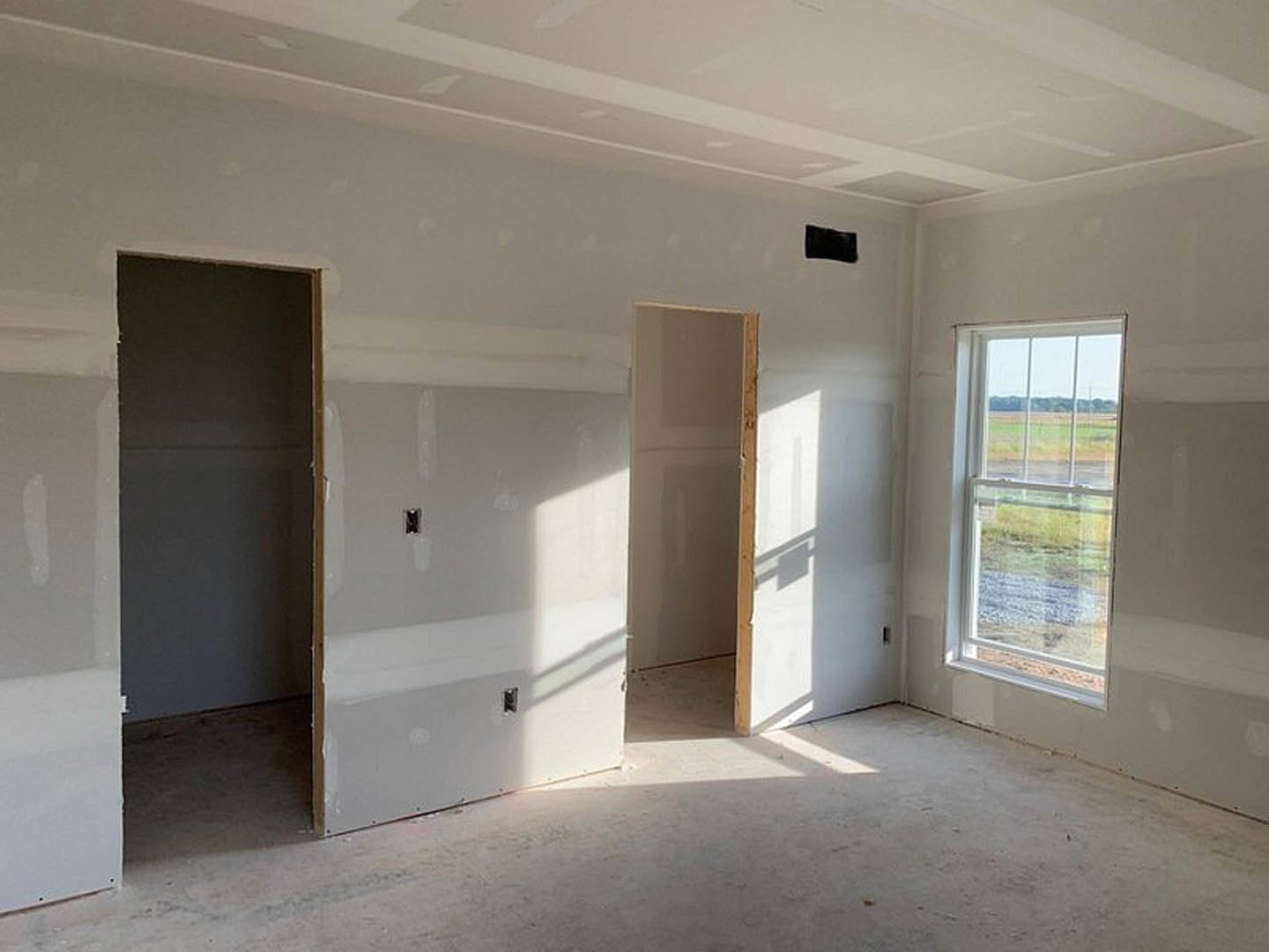 White-painted room with a closed door, rectangular window overlooking grass and fence, light-colored flooring, and smooth plaster walls.